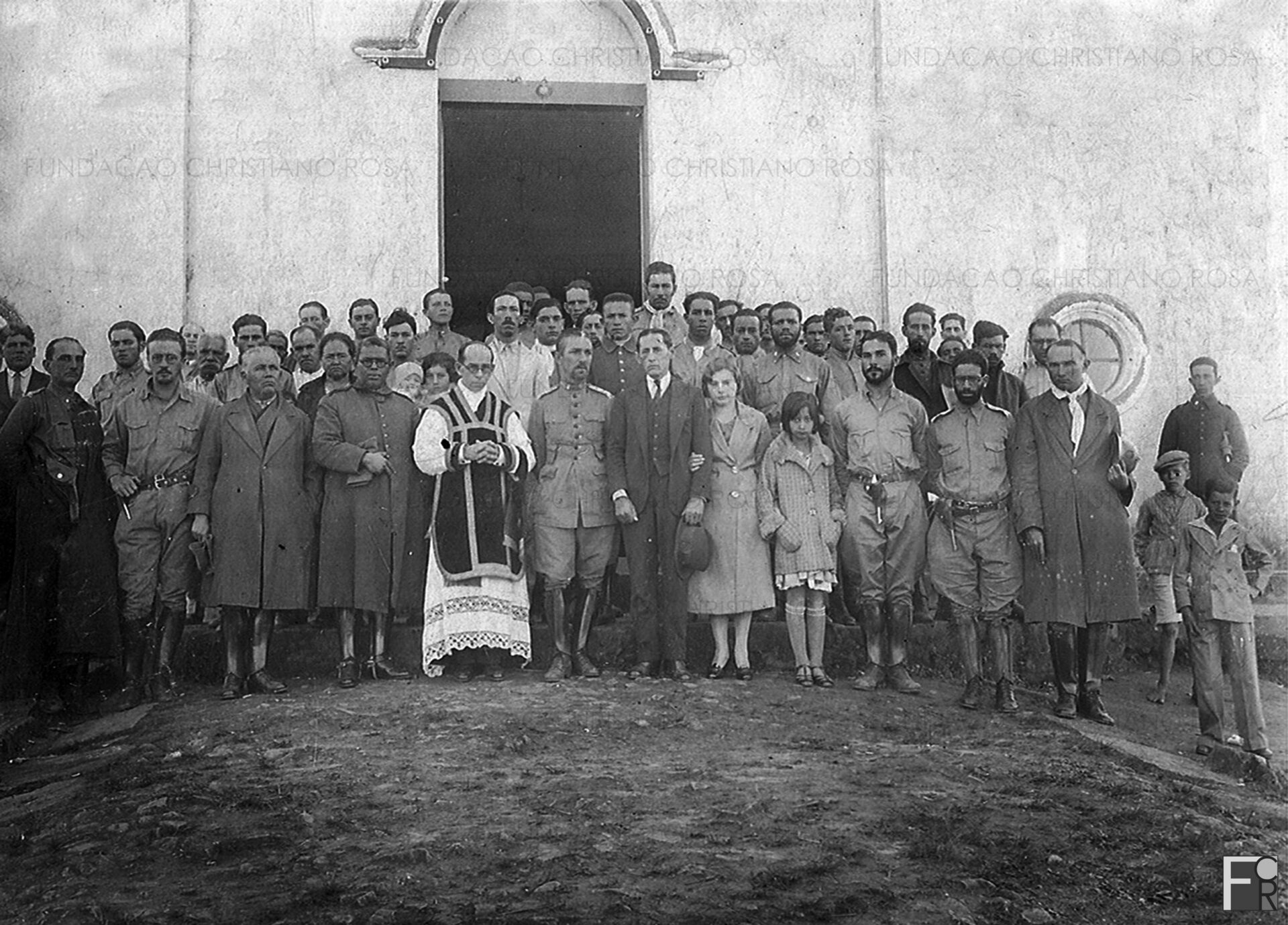Após missa celebrada na Matriz de São Miguel, em Piquete/SP, um registro fotográfico que mostra o comandante militar Leôncio Neiva ladeado pelo padre Estanislau Leme e pelo prefeito Antônio Pelúscio, de braço com a filha Zenaide, e Zeli Ferreira. À esquerda do padre, o tenente coronel Felisberto Antônio Fernandes Leal, diretor da F.P.s.F. entre agosto e setembro de 1932. Moradores da cidade reconhecidos: Joaquim Sacristão; D. Rita, esposa do sr. Juca Pinto, com a filha, Mocinha, que carrega uma criança ao colo; José Vieira Soares, Salviano e José Alves Beraldo.