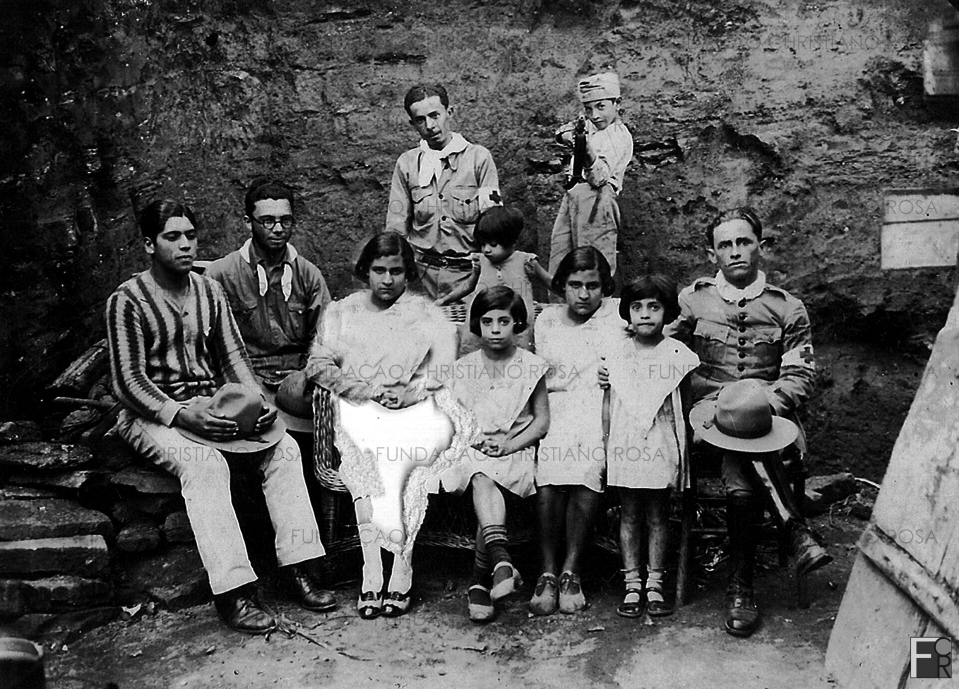 Três soldados do setor de saúde, instalado no Cine Glória, em Piquete, SP, com os filhos do senhor Benedito Elias: João, Geralda, Bibiu (perna cruzada), Lavínia (menor) e Orlando Siriri (com a arma), ?, ?. Foto do acervo da Fundação Christiano Rosa, de Piquete, SP.